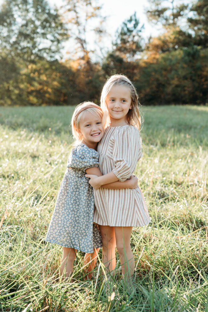 Two sister is huging and smiling in the camera while standing in the grass field in Birmingham Park, Atlanta during thier family photo session.