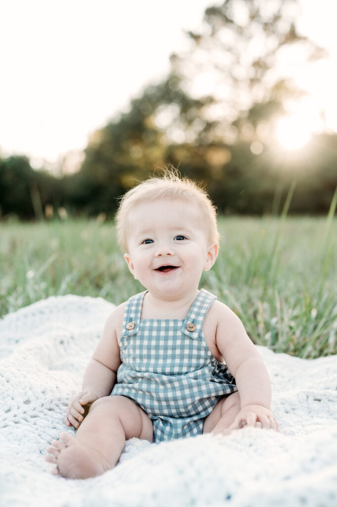 Little brother is sitting on a blanket in the grass and smiling during a family session in Birmingham Park, Atlanta