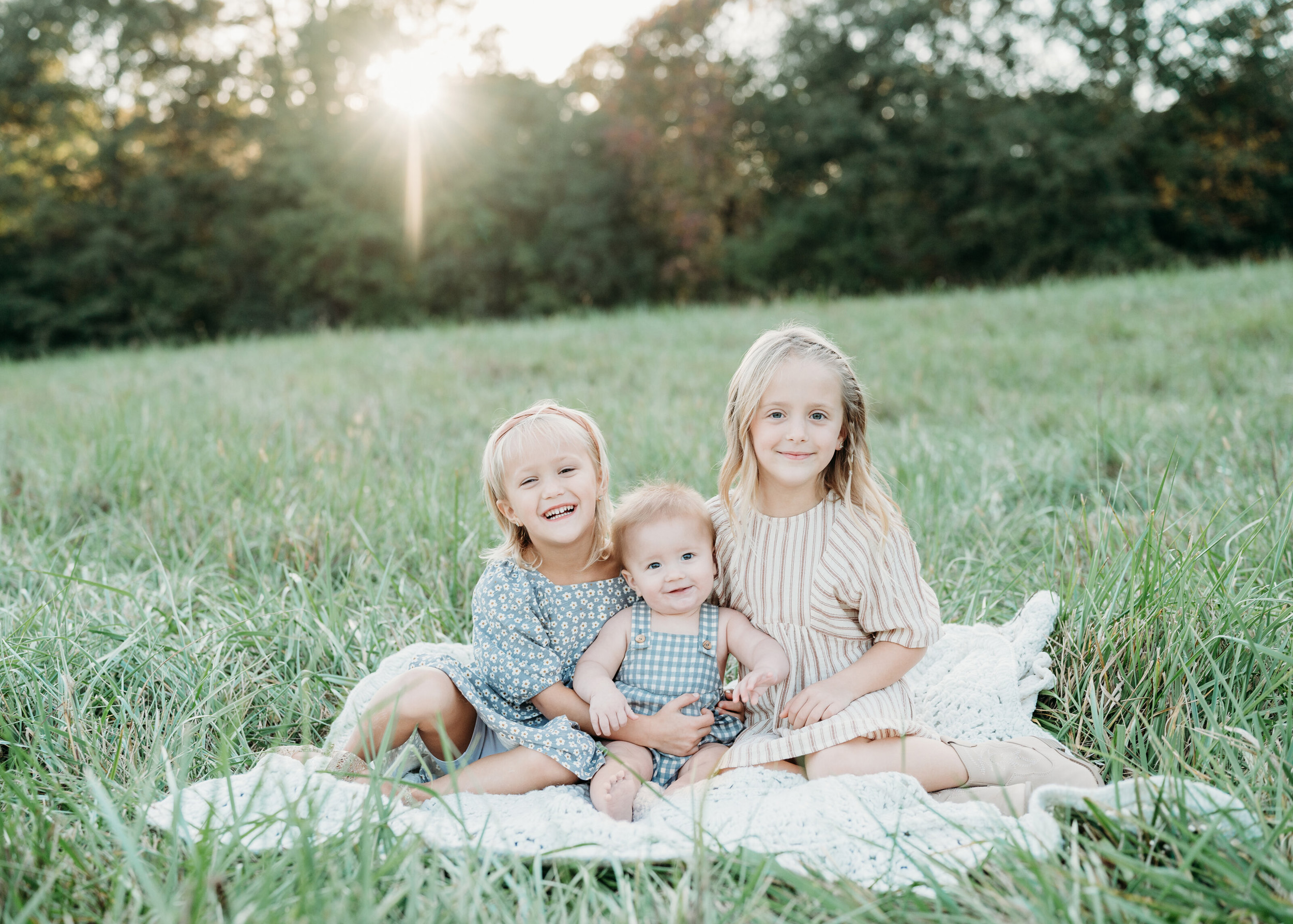 Three siblings are sitting on a blanket in the grass and smiling during a family session in Birmingham Park, Atlanta