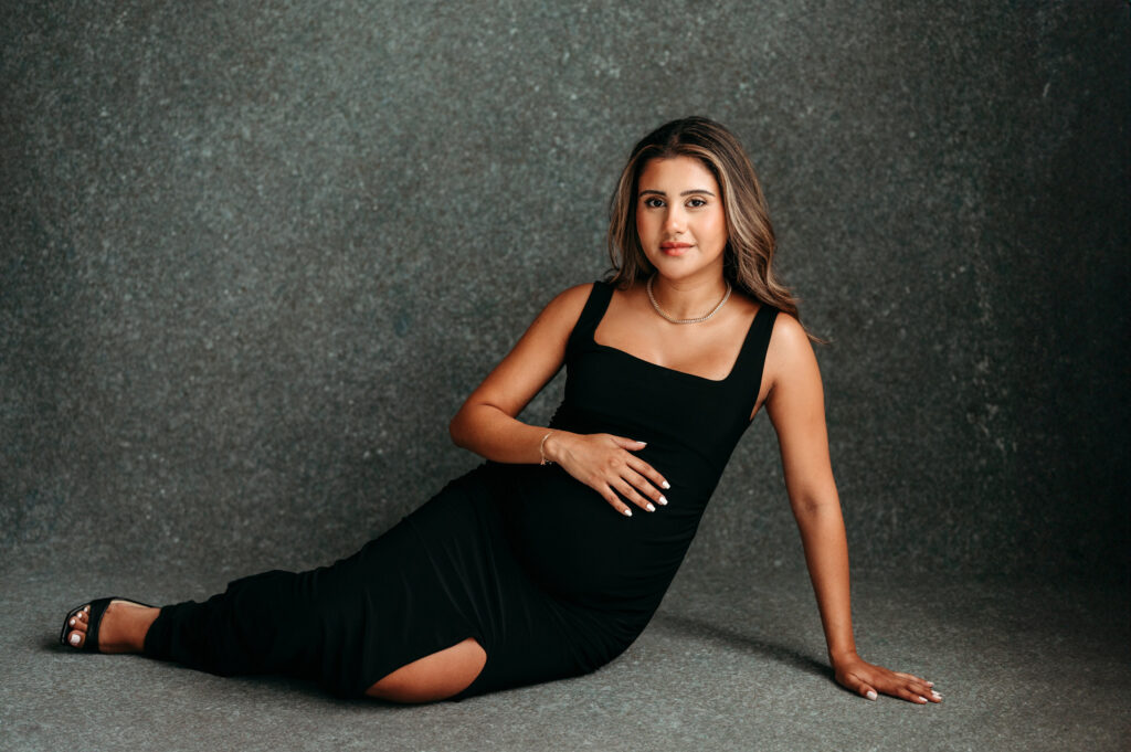 Pregnant mama in black dress sitting down holding her hand over her belly in her studio maternity session in North Atlanta