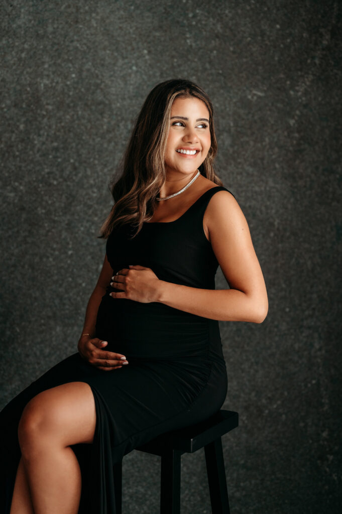 Pregnant woman sitting in a blakc dress looking over her shoulder in her studio maternity session in Atlanta